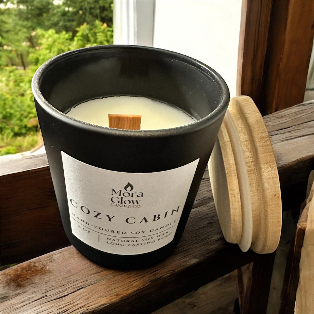 8 oz. Cozy Cabin soy candle sitting in window, black jar with bamboo lid and a wooden wick, find your calm, embrace the glow.